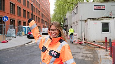 Junge ZÜBLIN Mitarbeiterin mit PSA vor einer Baustelle.