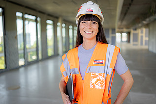 Foto von einer lachenden ZÜBLIN-Architektin in Arbeitssicherheitskleidung auf einer Baustelle mit Tablet im Arm.