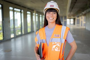 Foto von einer lachenden ZÜBLIN-Architektin in Arbeitssicherheitskleidung auf einer Baustelle mit Tablet im Arm.