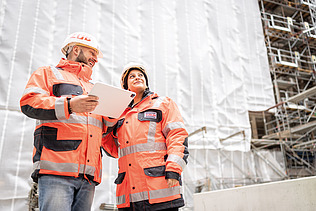Two employees wearing high-visibility clothing are talking on a building site; one is holding a tablet