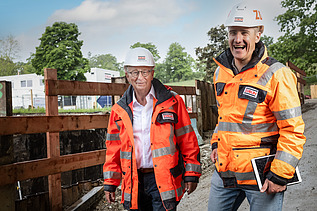 Zwei Mitarbeiter in Arbeitsschutzkleidung auf der Baustelle, lachen in die Kamera