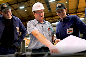Drei Mitarbeiter in einer Werkstatt, der Mann in der Mitte zeigt auf einen Plan.