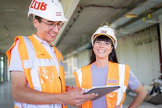 Foto von zwei ZÜBLIN-Mitarbeitenden, die lachend zusammen auf der Baustelle stehen und etwas besprechen.