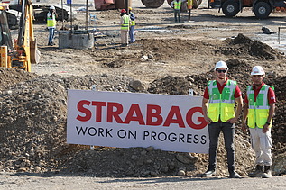 Zwei Mitarbeiter in Arbeitsschutzkleidung auf einer Baustelle, im Hintergrund ein STRABAG-Banner