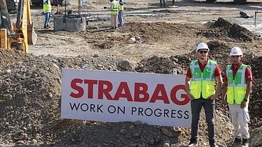 Zwei Mitarbeiter in Arbeitsschutzkleidung auf einer Baustelle, im Hintergrund ein STRABAG-Banner