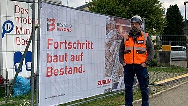 Oberpolier Mike in Arbeitskleidung vor einem Bauzaun mit Aufschrift 