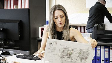 Foto von einer Frau, die am Schreibtisch sitzt und auf einen Bauplan schaut, während im Hintergrund ein weiterer Mitarbeiter arbeitet