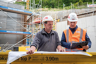 Two men in high-visibility workwear, one holding a plan, the other holding a tablet