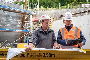 Two men in high-visibility workwear, one holding a plan, the other holding a tablet