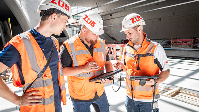 Three men in high-visibility workwear are discussing a building plan.
