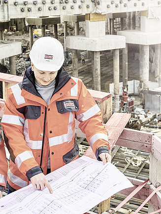Junge Frau und junger Mann in Arbeitskleidung auf einer Baustelle, zeigen auf Bauplan