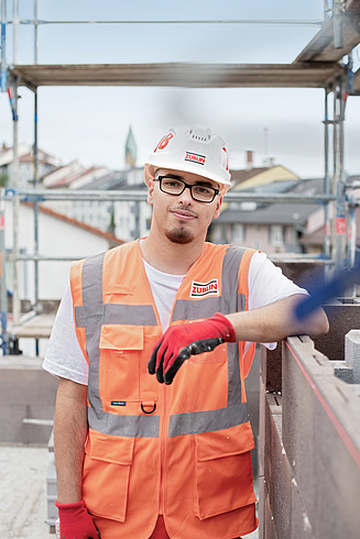 A young apprentice is standing on a building site wearing a high-visibility vest and a hard hat.