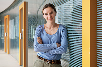 Junge Frau mit verschränkten Armen vor einer Glaswand, lächelt in die Kamera