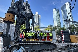 Kinder in Arbeitsschutzkleidung stehen auf einer Baustelle auf einem Riesenbohrgerät, im Hintergrund die Frankfurter Skyline