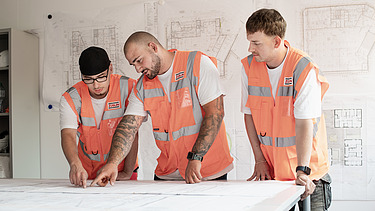 Three young men are standing in the site office in front of a building plan.