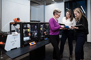 Drei ZÜBLIN Mitarbeiterinnen, die vor einem 3D Drucker stehen und etwas besprechen.