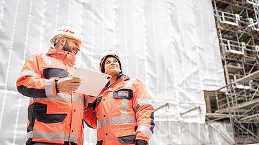 Zwei Mitarbeitende in Arbeitsschutzkleidung auf einer Baustelle im Gespräch, einer mit Tablet