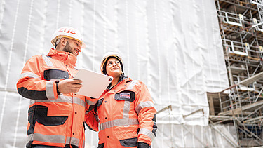 Two employees wearing high-visibility clothing are talking on a building site; one is holding a tablet