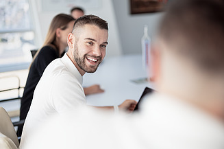 A man is sitting in a meeting, smiling, and turns to a colleague.