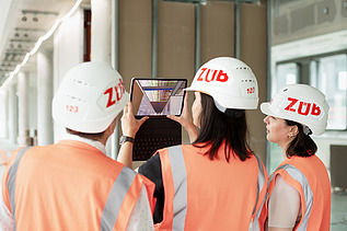 Photo of three ZÜBLIN staff members on a building site, viewing a digital building plan on a tablet.