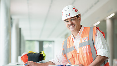 A photograph of a ZÜBLIN employee standing on a building site, poring over a construction plan and smiling at the camera.