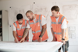 Three young men are standing in the site office in front of a building plan.