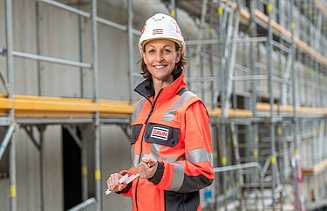 Frau steht mit persönlicher Schutzausrüstung auf einer Baustelle und lächelt in die Kamera.