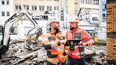 Zwei Mitarbeitende in Arbeitsschutzkleidung auf einer Baustelle im Gespräch, einer mit Tablet