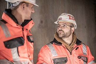 Interview Alex ZÜBLIN Mitarbeiter mit Bauhelm guckt einen weiteren Mitarbeiter an.