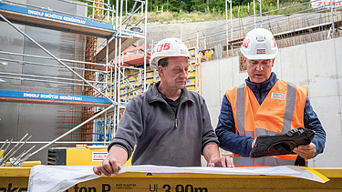 Two men in high-visibility workwear, one holding a plan, the other holding a tablet