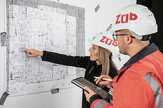 Two staff members wearing hard hats are looking at a building plan.