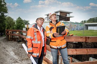 Zwei Mitarbeiter in Arbeitsschutzkleidung auf der Baustelle mit Tablet und Bauplan in der Hand