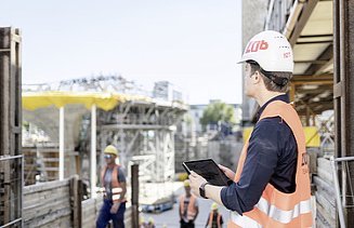 Foto von einem Mann mit Helm, Sicherheitsweste und Tablet, der auf eine Baustelle schaut