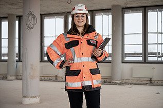 Foto einer ZÜBLIN-Mitarbeiterin, die in Arbeitsschutzkleidung und einem Tablet auf einer Baustelle steht und in die Kamera schaut.