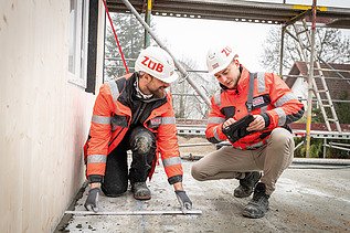 Zwei Mitarbeitende in Arbeitsschutzkleidung auf einer Baustelle im Gespräch, einer mit Tablet, einer mit Zollstock