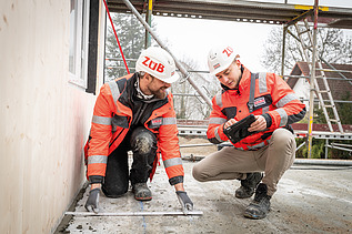 Two staff members in high-visibility clothing talking on a building site; one is holding a tablet, the other a folding rule