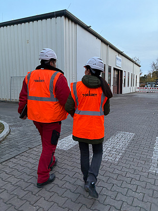 Zwei Männer in Arbeitsschutzkleidung gehen über eine Baustelle, im Hintergrund ist eine Lagerhalle zu sehen