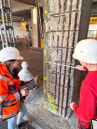 Zwei Mitarbeitende in Arbeitssicherheitskleidung, eine links mit Tablet, der andere rechts mit Zollstock