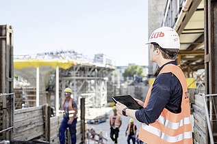 Foto von einem Mann mit Helm, Sicherheitsweste und Tablet, der auf eine Baustelle schaut